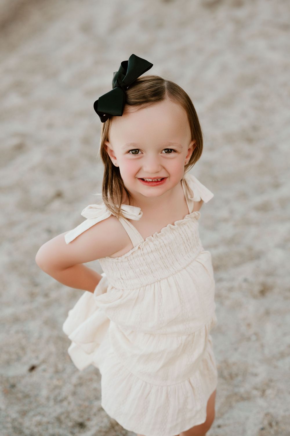A small child wearing a white ruffled dress and black bow twirls and smiles during an outdoor photo session.