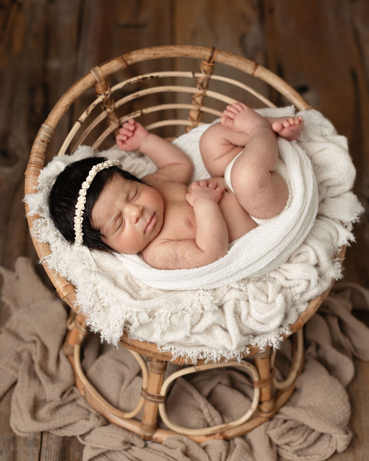 A sleeping newborn baby rests peacefully in a white woven basket on soft brown fabric. Elizabeth Lang Photography Surrey