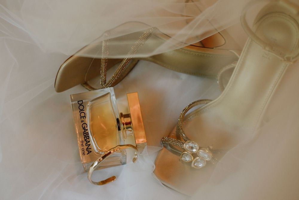 Flat lay image of the bride’s shoes, perfume, and gold jewellery with the veil softly draped over them, capturing elegant wedding details