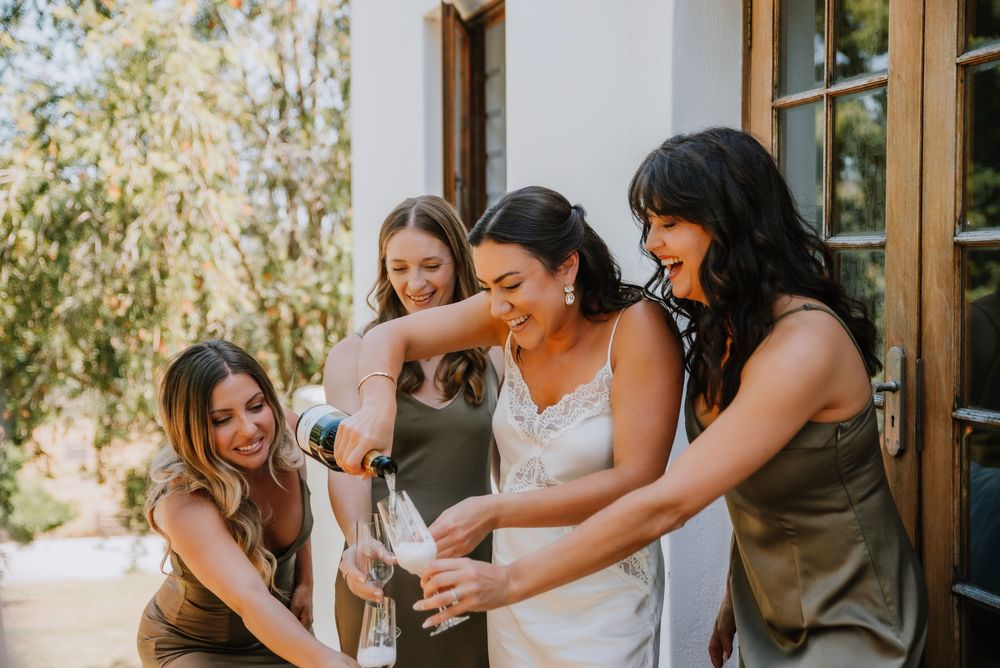 Cheerful wedding moment as the bride pours champagne into bridesmaids’ glasses after popping the bottle, capturing a joyful celebration