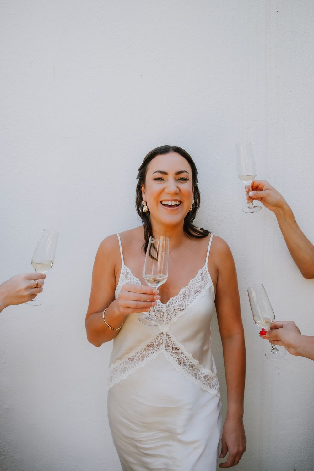Quirky image of the Bride with a big beautiful smile on her face.  In fornt of a white wall surrounded by champagne glasses