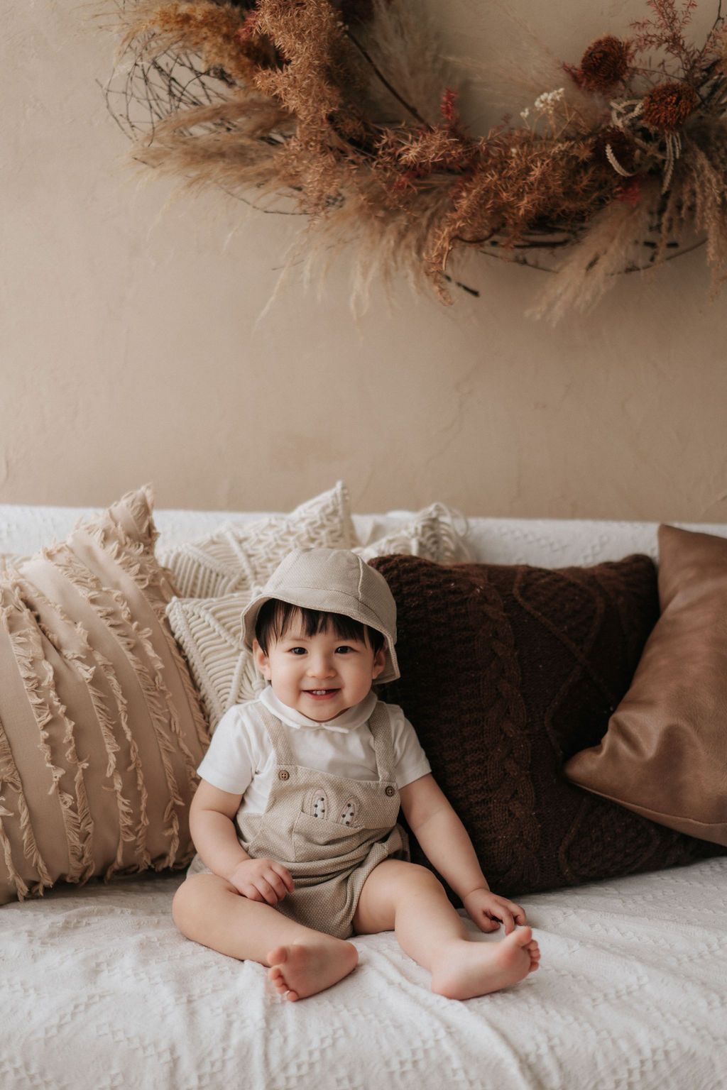 Baby wearing a white hat sits on white bedding with brown throw pillows in a series of candid shots.