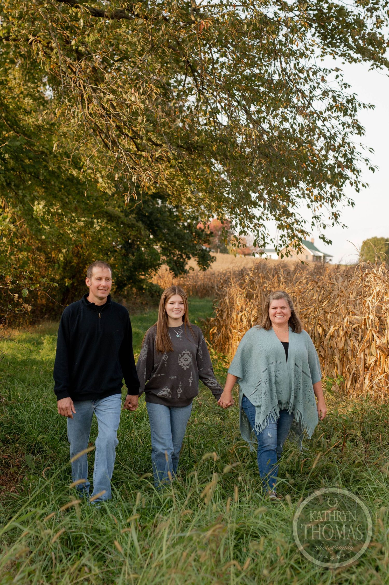 Three people walking together through tall grass with autumn trees and cornfield in background.