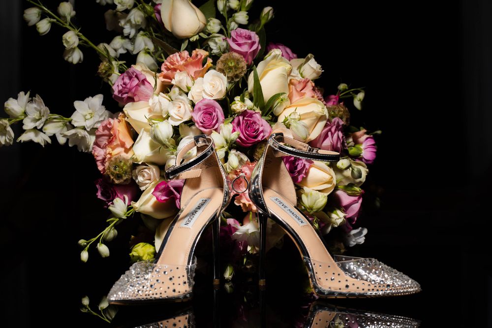Sparkly bridal shoes arranged with pink and cream roses against dark background.