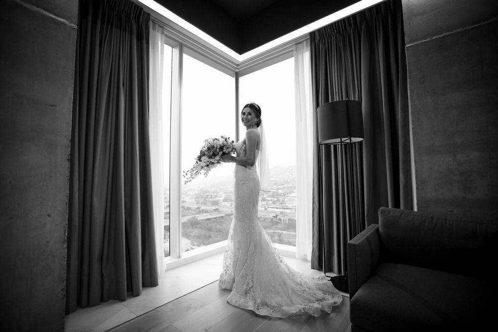 Black and white photo of bride holding bouquet by window in luxury hotel room.