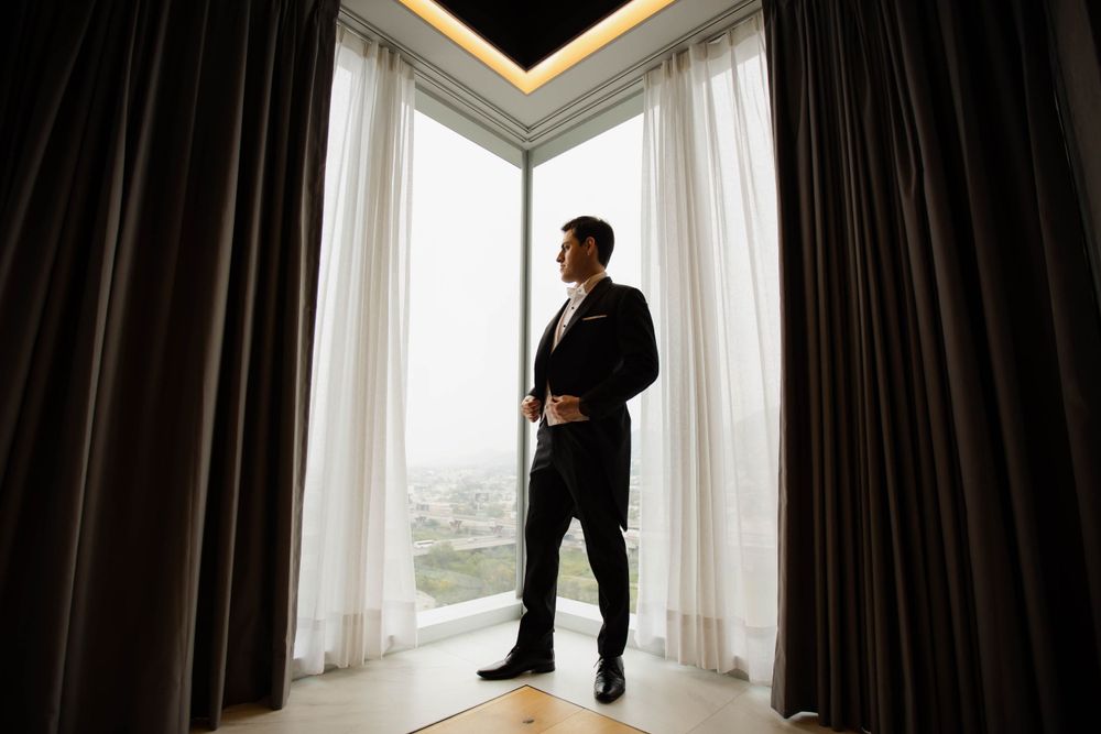 Silhouette of groom standing by tall windows in modern room with dramatic lighting.