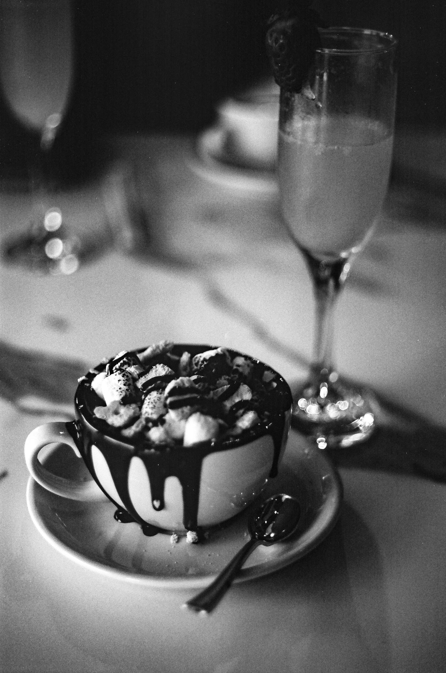 Black and white photo of a decadent hot chocolate topped with whipped cream and chocolate drizzle beside a champagne flute.