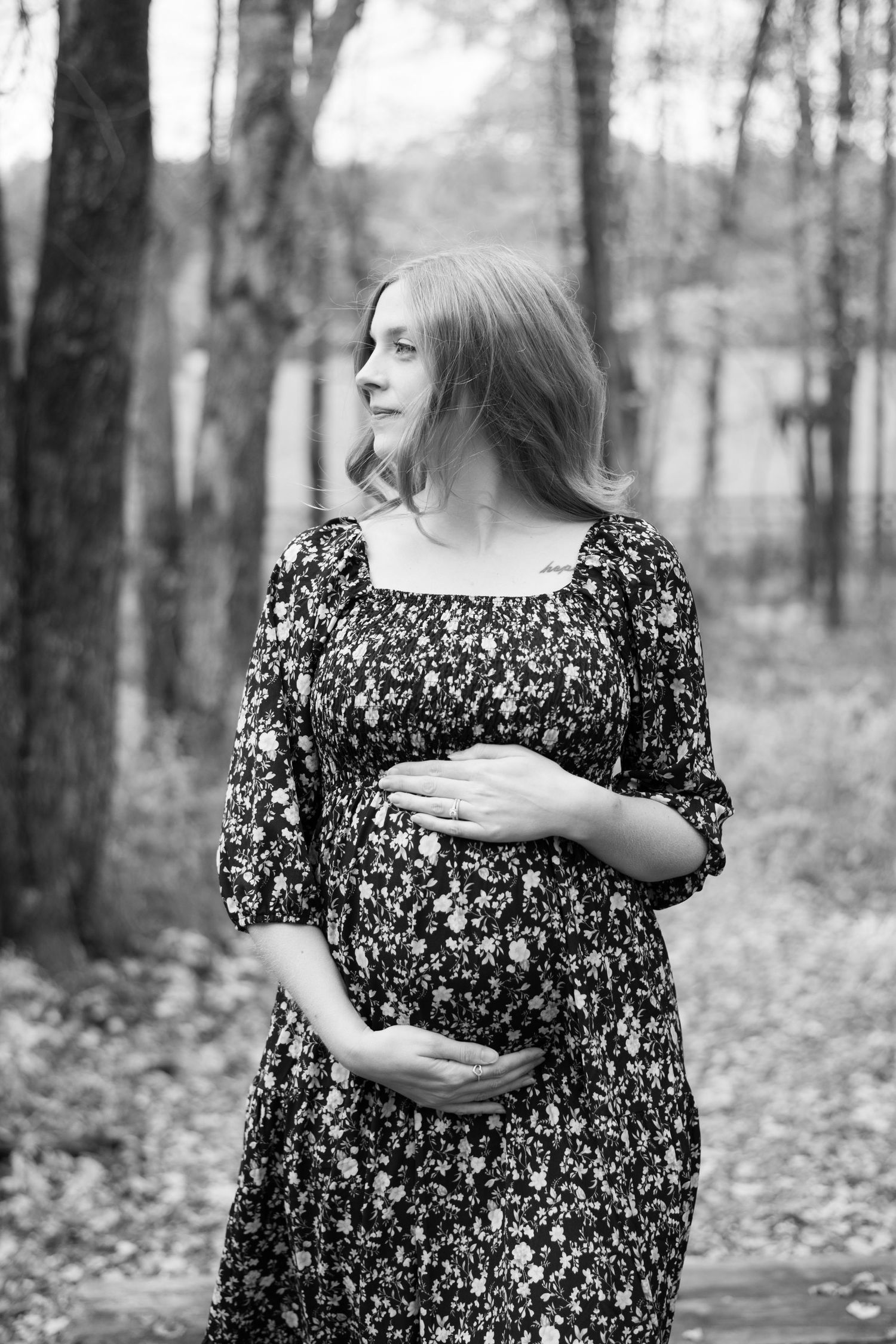 Black and white photo of expectant mother in floral dress standing in a forest setting.