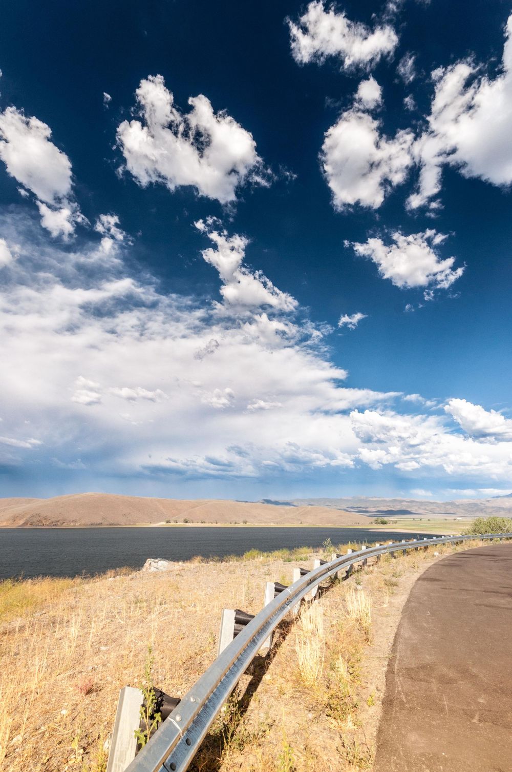 Winding guardrail along a desert road with dramatic blue sky and white clouds above.