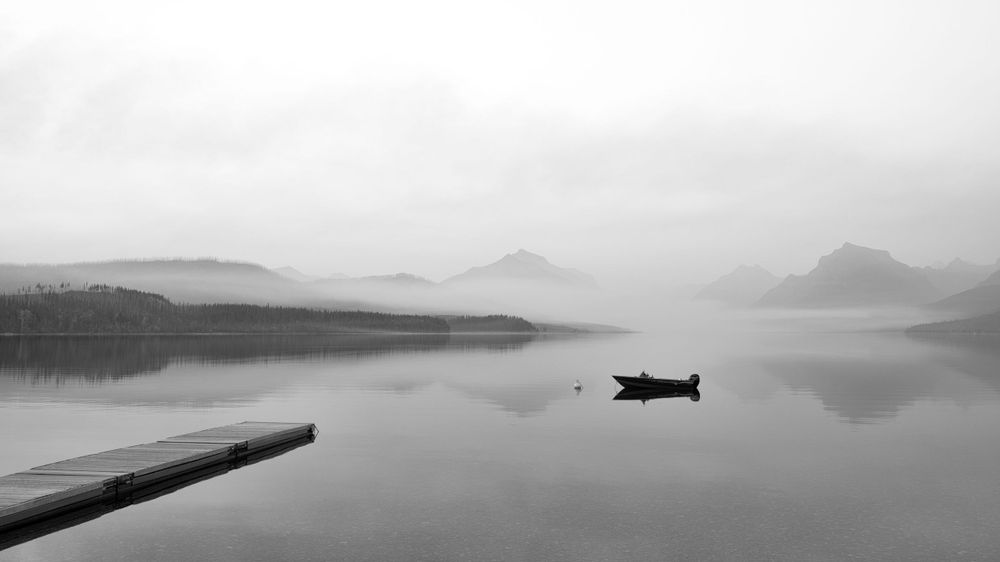 Minimalist black and white landscape of a small boat on a misty lake surrounded by mountains.