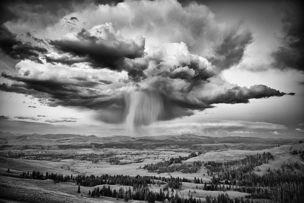 Dramatic storm clouds with rain over a vast mountain landscape in black and white.