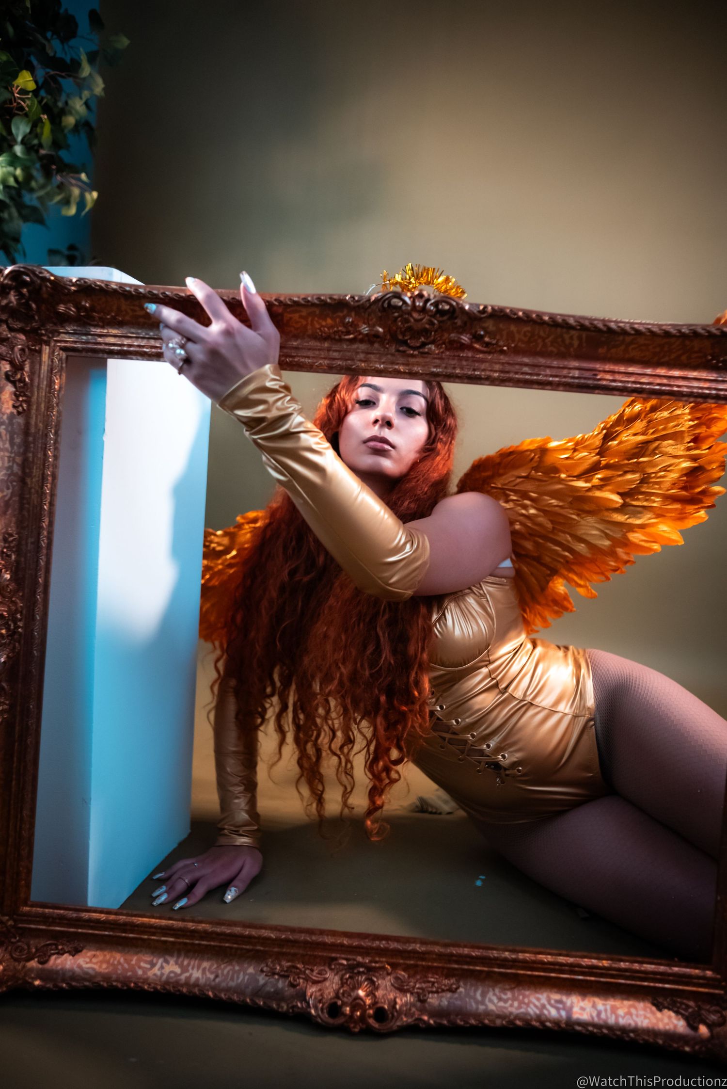 Artistic portrait in gold tones featuring large decorative wings against an ornate frame with dramatic lighting.