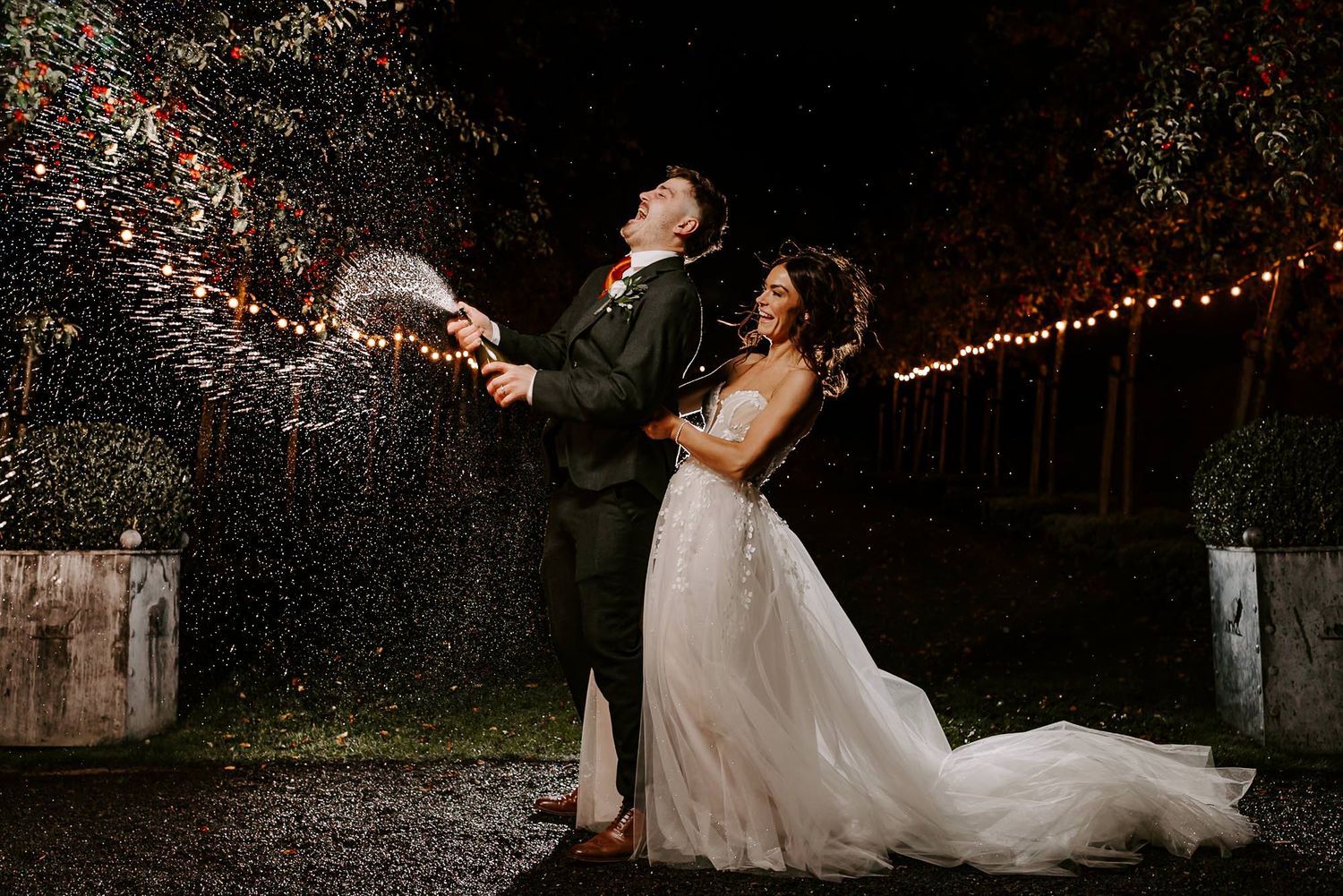 A couple in formal wedding attire dances and twirls in a romantic outdoor setting with twinkling lights.