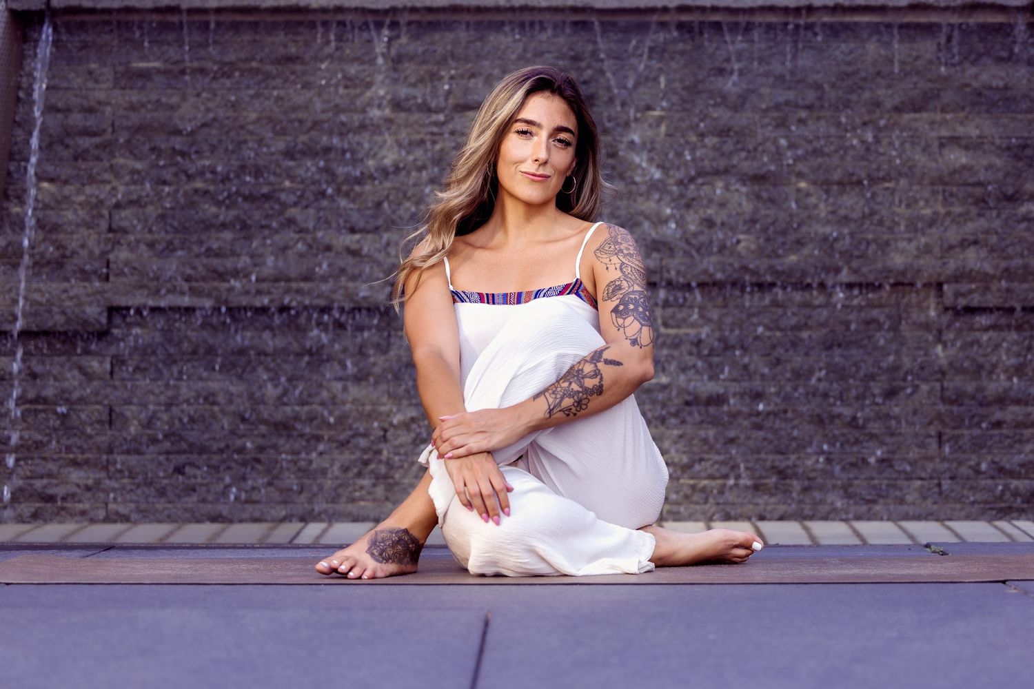 A person in a white dress sits on the ground against a gray wall, showing colorful tattoos on their arms.