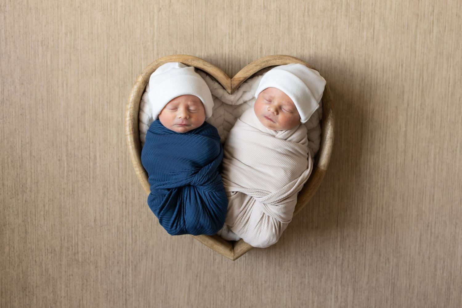 A swaddled newborn lies in a heart-shaped basket lined with pink fabric on a dark wooden surface.