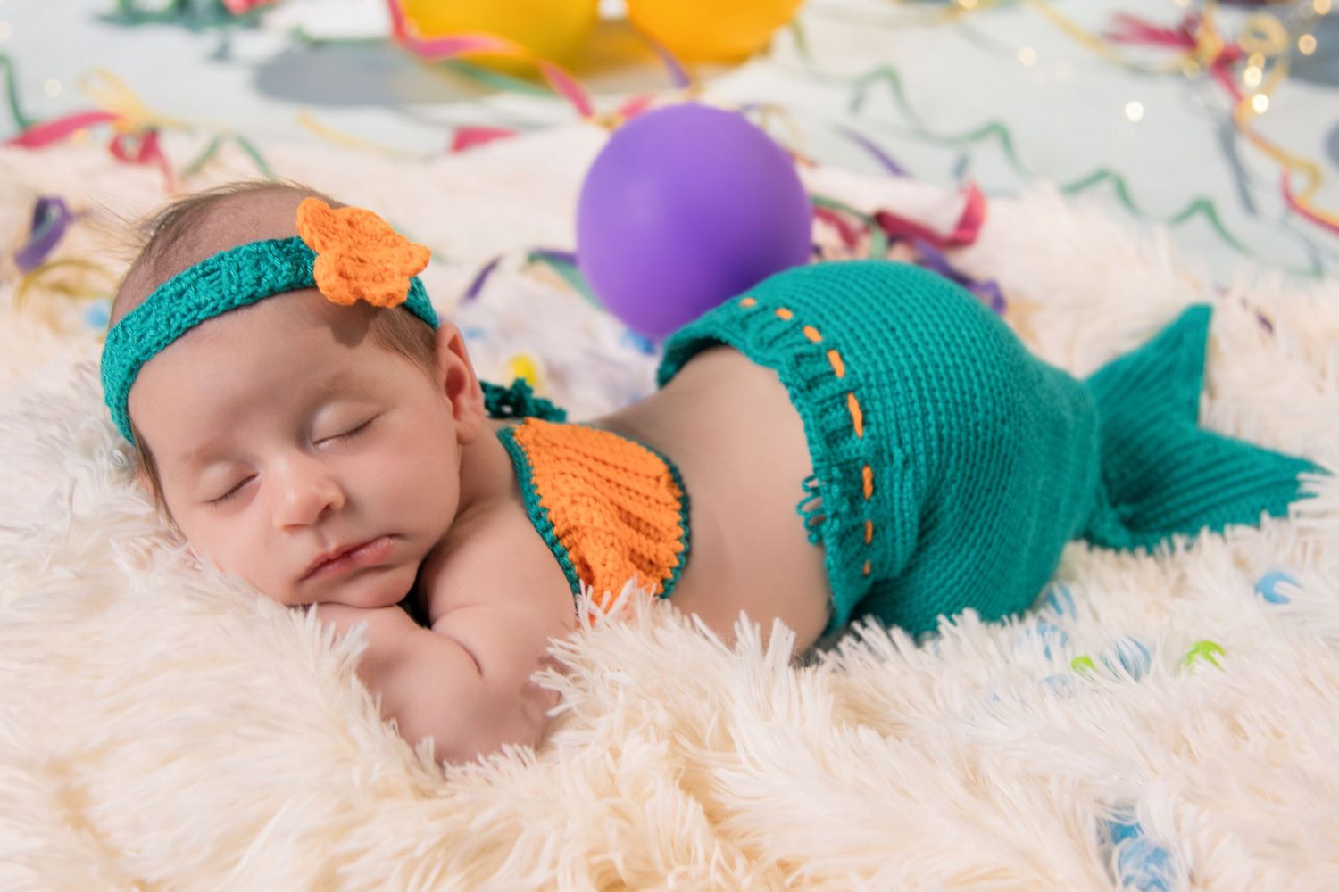 A sleeping baby wearing a teal outfit lies on white fur surrounded by colorful butterflies and balloons.