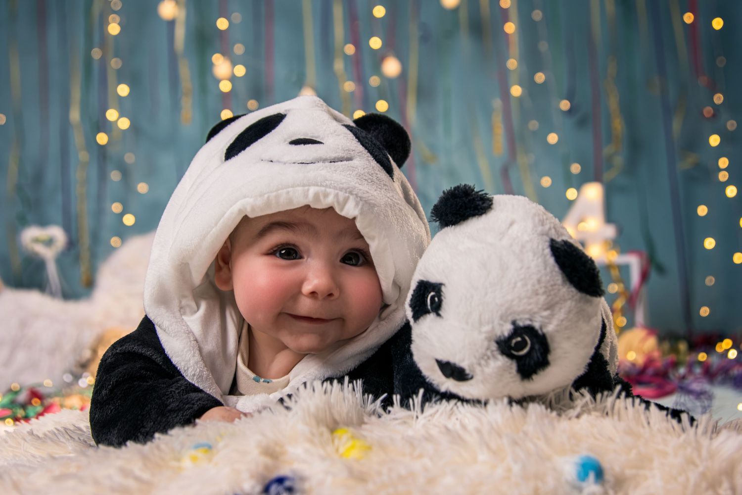 A baby in a panda costume lies on white fur with twinkling lights in the background.