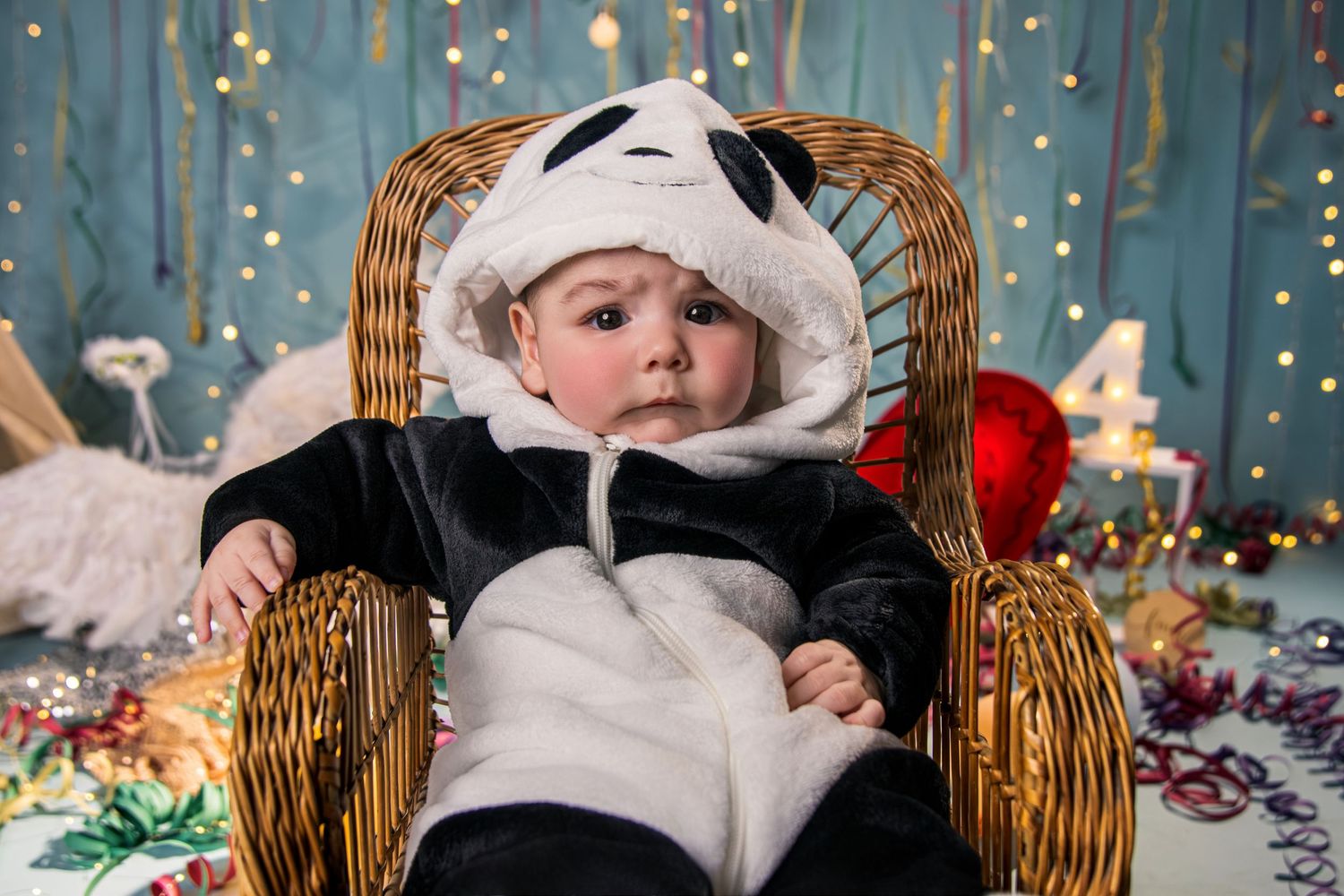 A baby wearing a white furry winter outfit sits in a wicker basket surrounded by twinkling holiday lights.