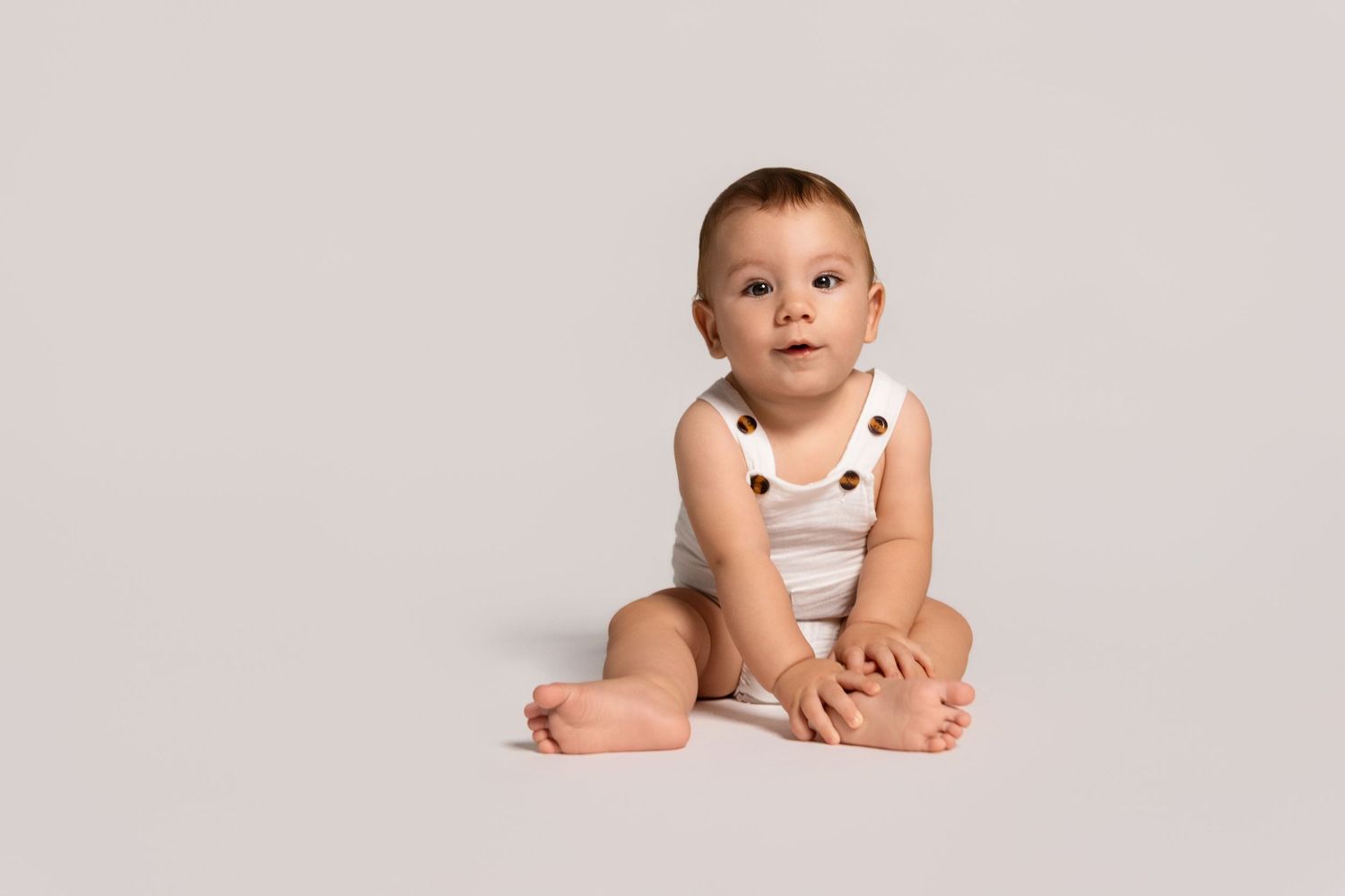 Baby in white outfit lying on light background during studio photoshoot.