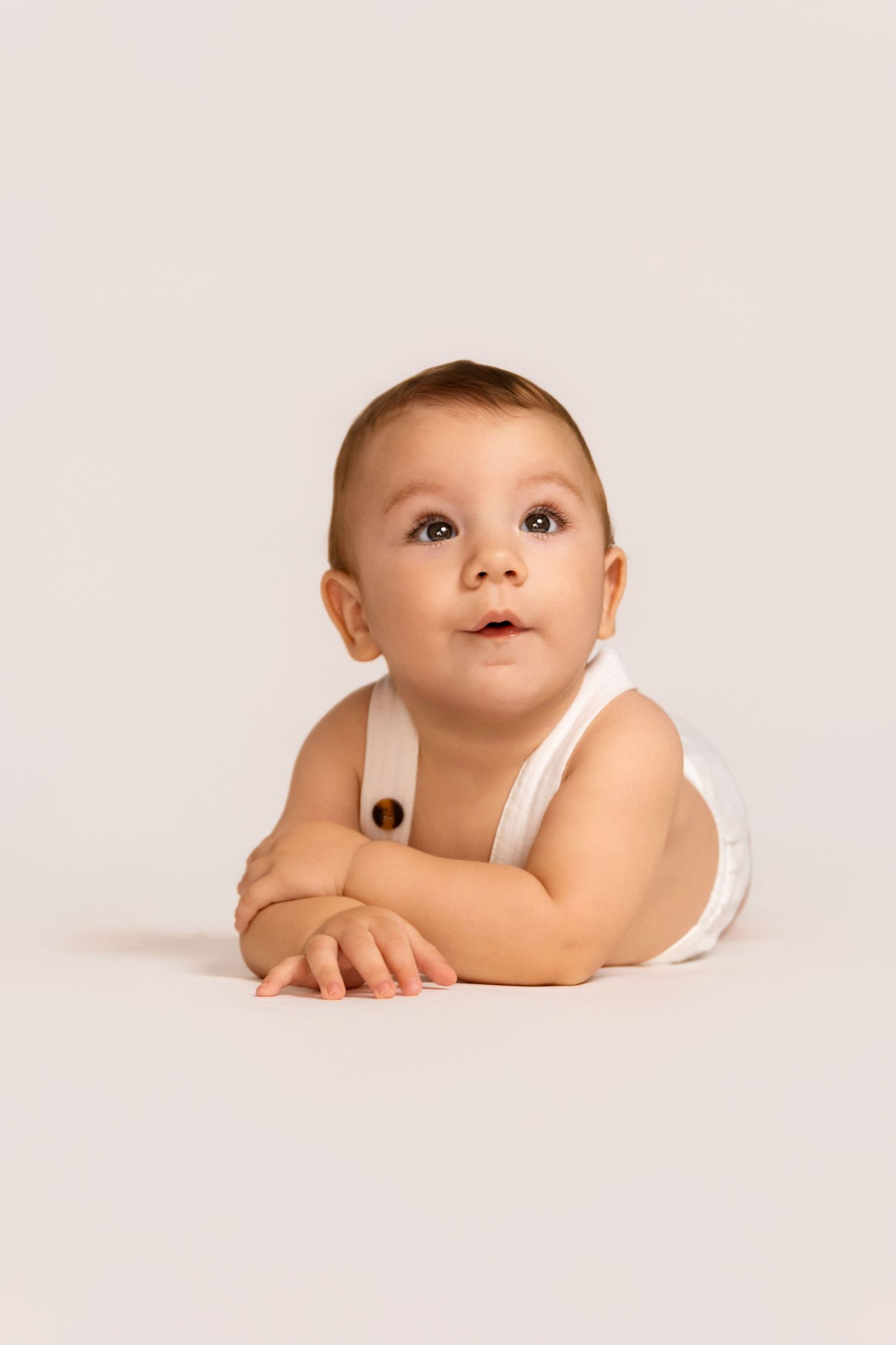 Baby in white outfit lying on light background during studio photoshoot.