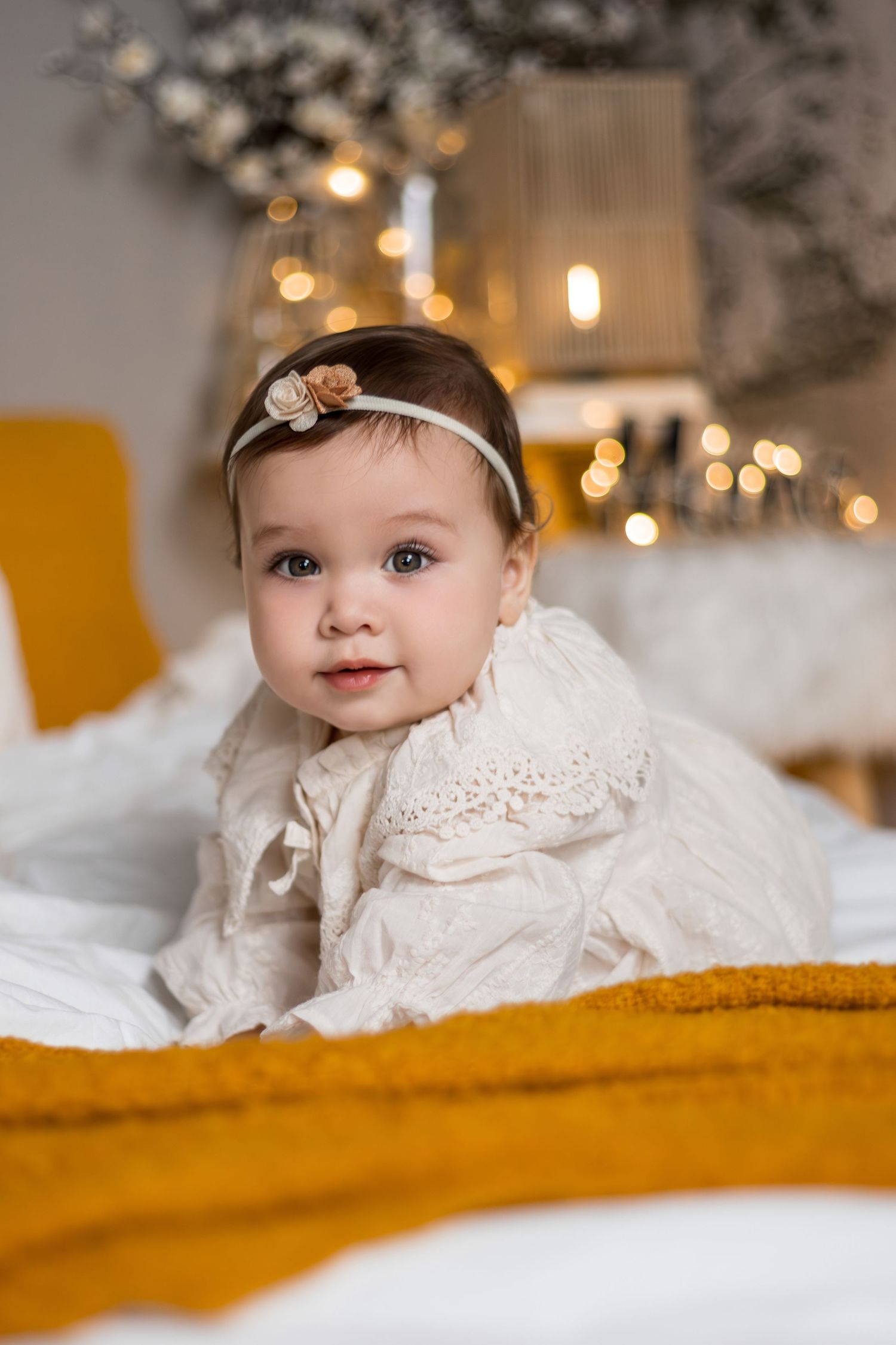 Baby in vintage white lace dress and headband lying on yellow blanket with twinkling lights in background.