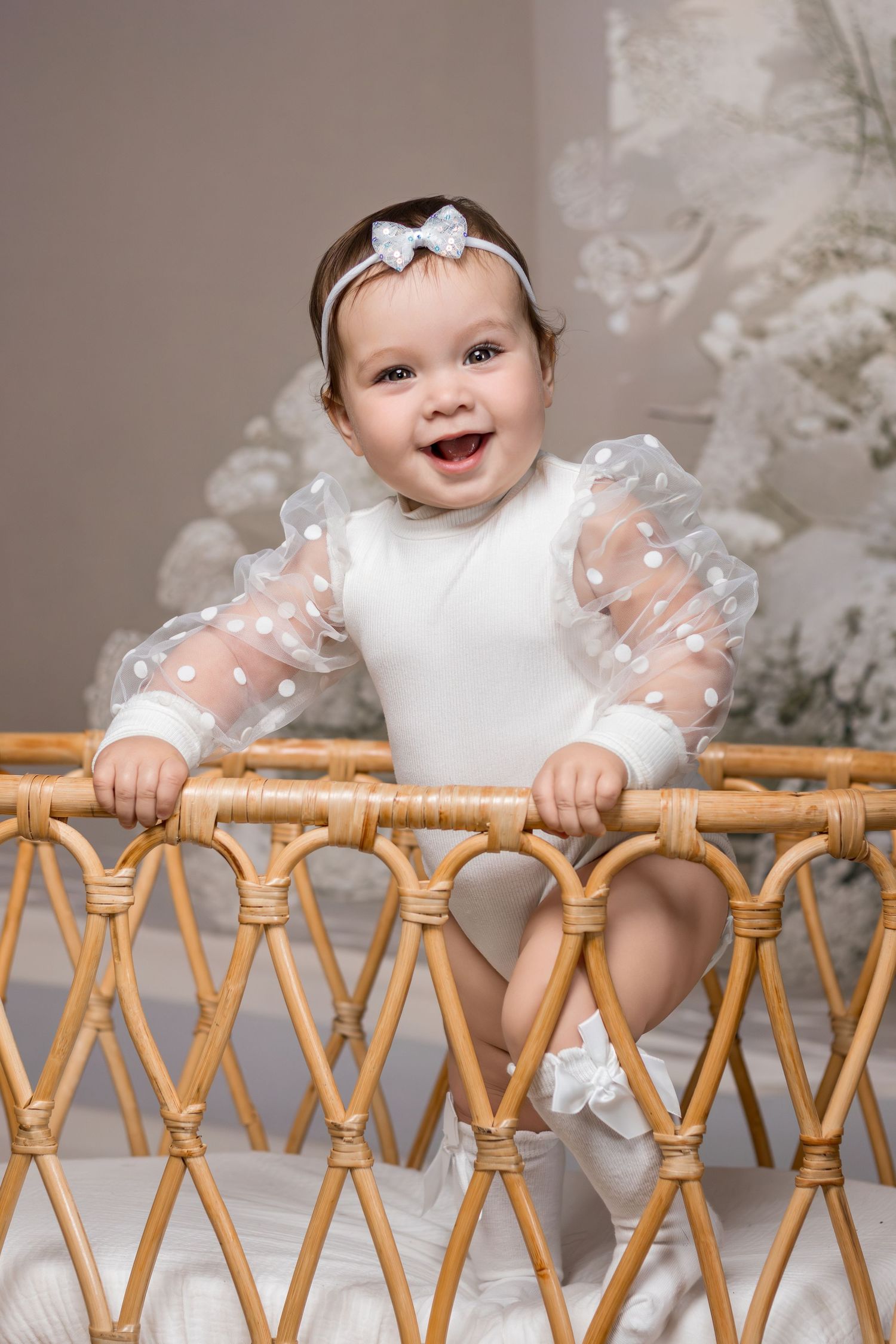 Baby in white lace dress with bow headband smiles while standing at rattan furniture during portrait session.