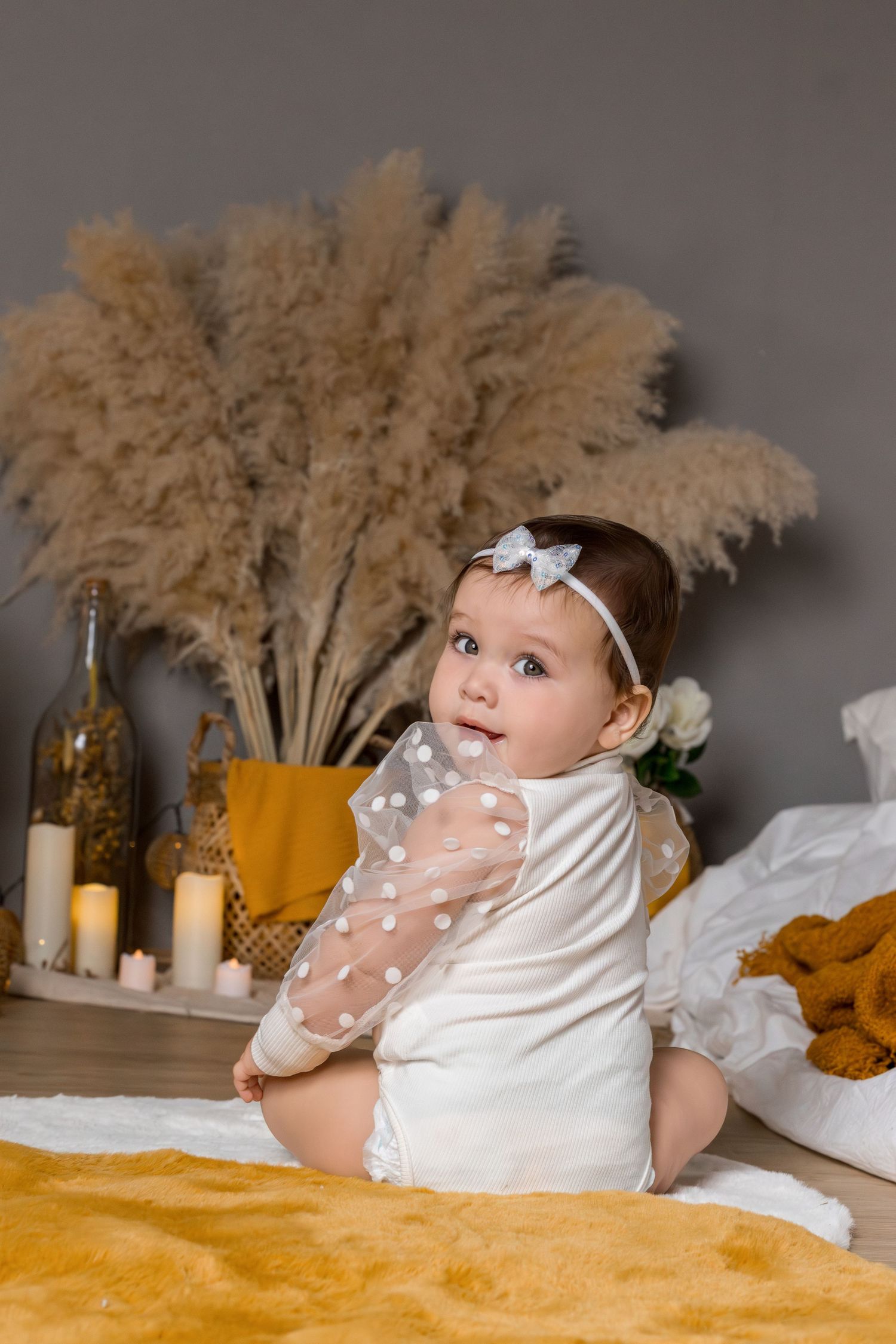 A baby in white dress lying on yellow bedding with dried pampas grass decor in the background.