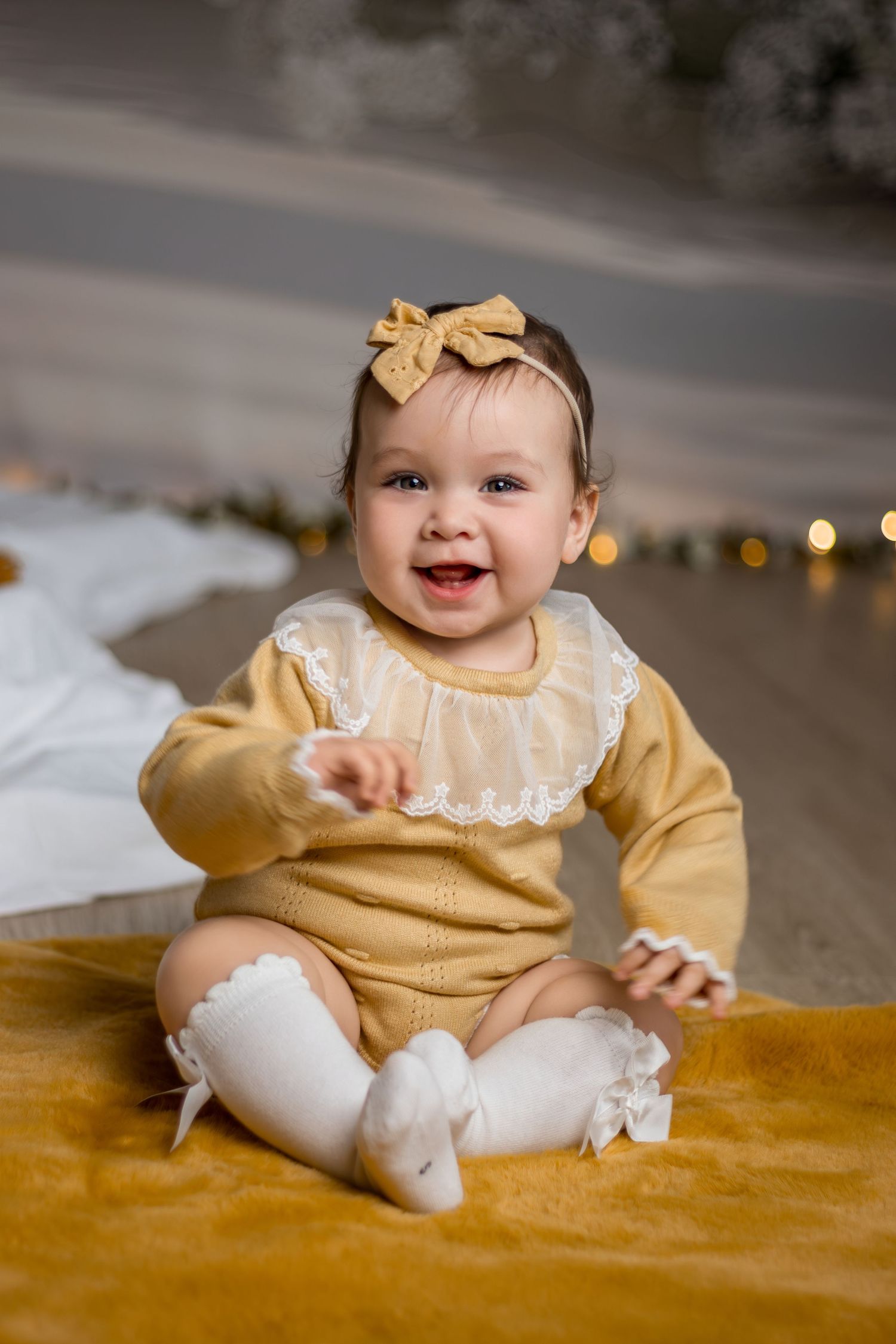 Baby in yellow and white dress sitting on mustard velvet blanket with a joyful expression.