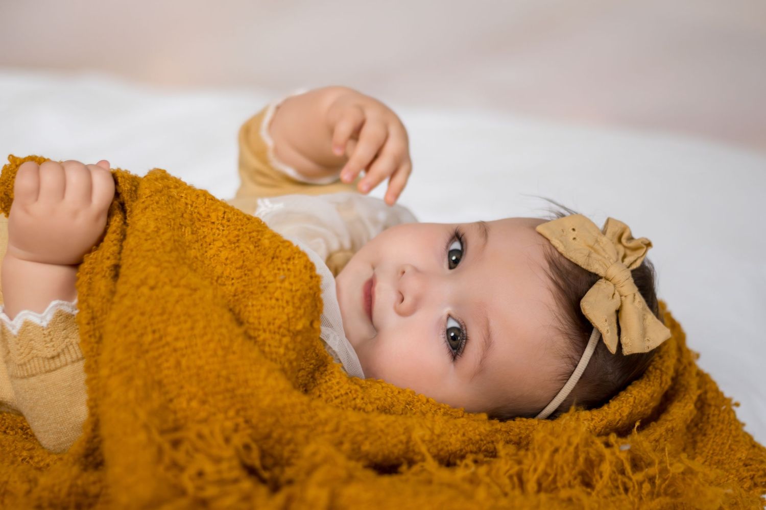 A child wearing a mustard yellow sweater and tan bow headband lies on a white surface.