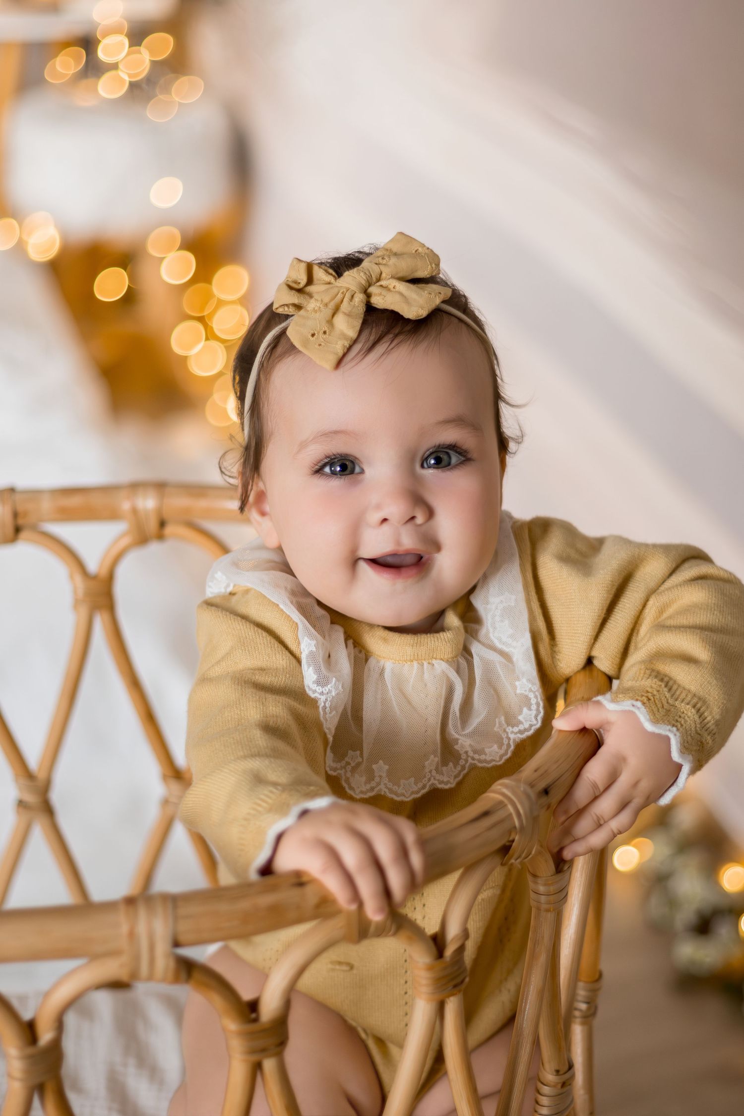 Baby in mustard yellow outfit with bow headband sits in rattan chair with twinkling holiday lights in background.