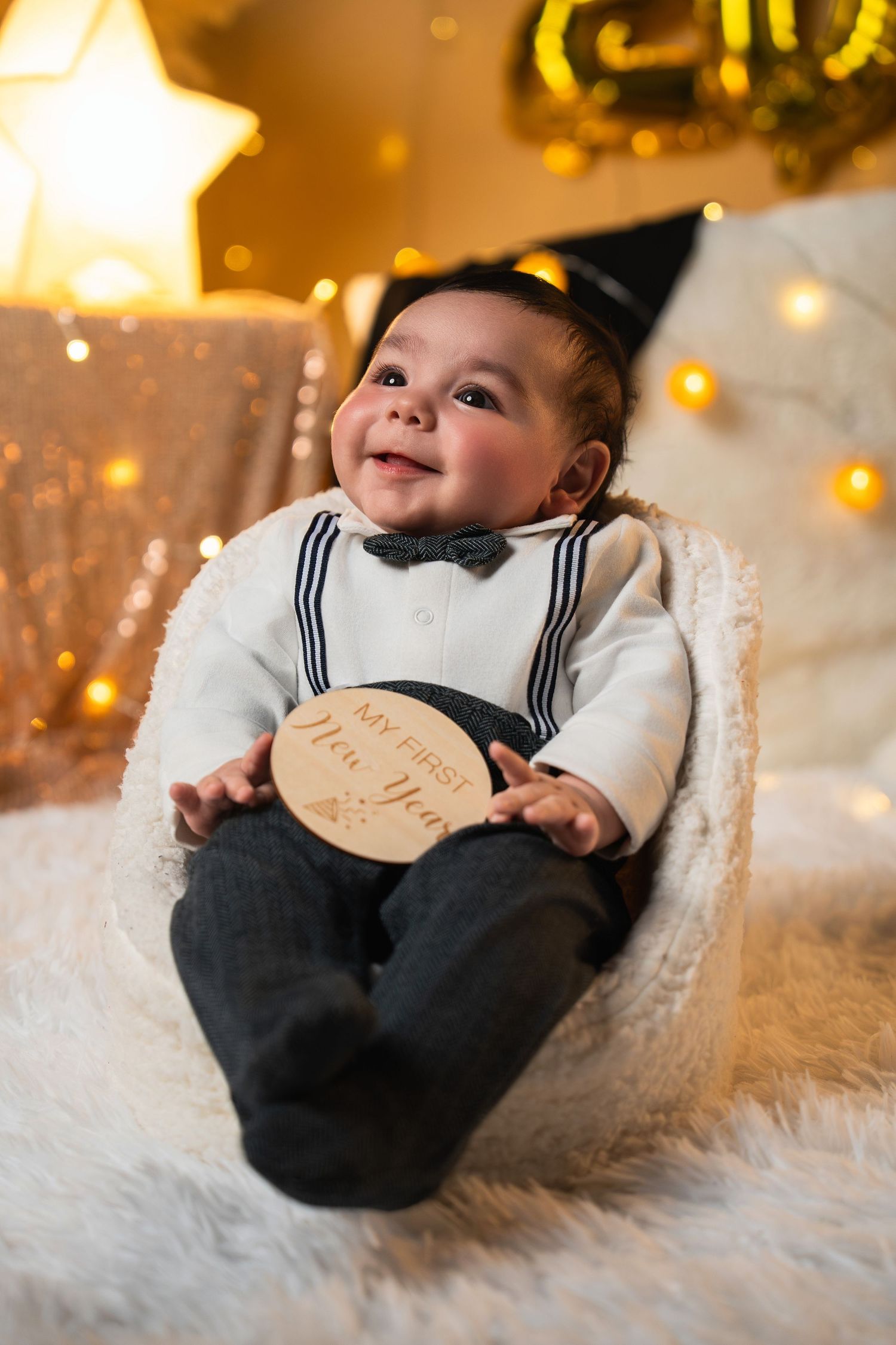 A baby in a navy sailor outfit smiles while lying on white fur with twinkling lights in the background.