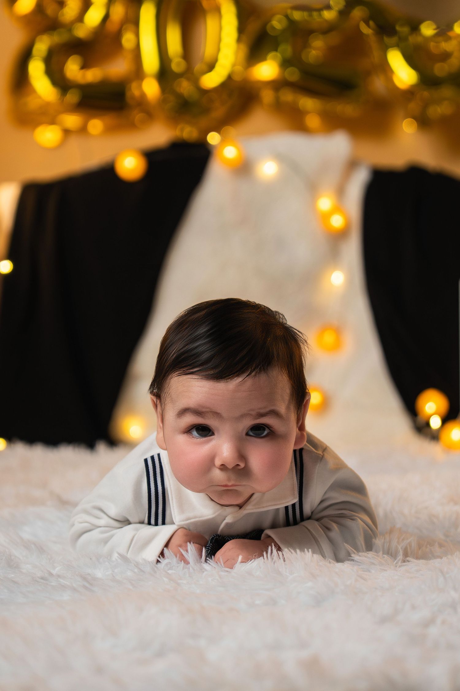 Baby lying on soft white blanket with warm glowing Christmas lights twinkling in background.