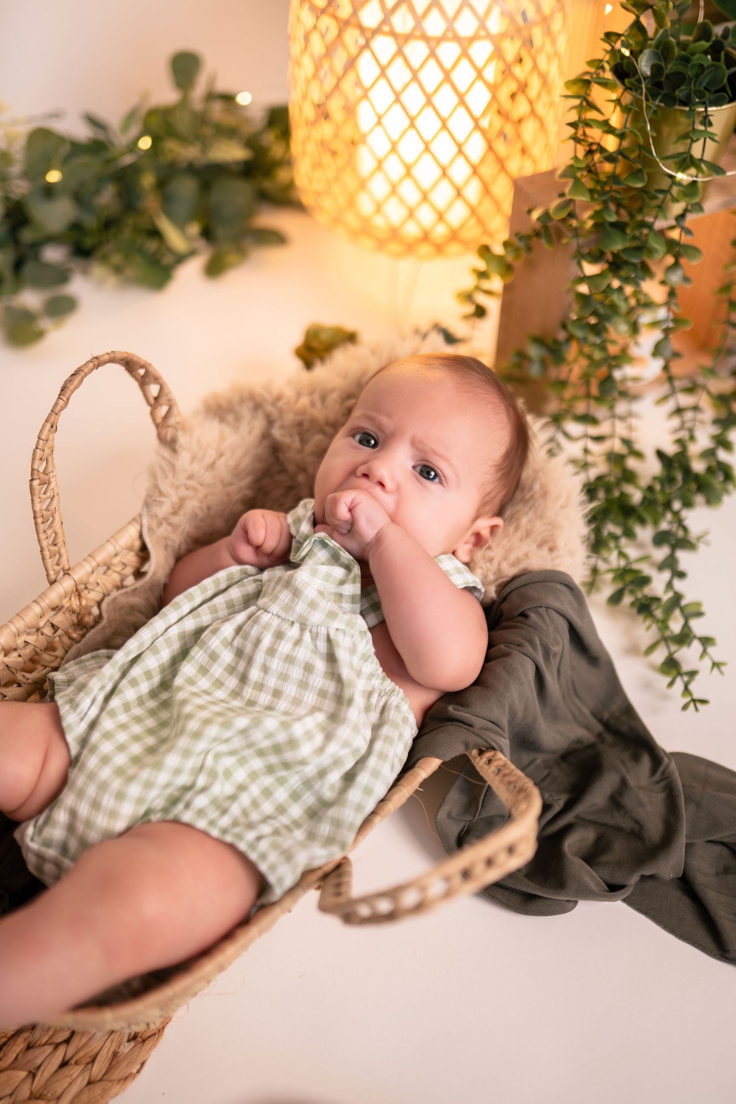 Infant lounging near decorative lamp with woven pattern and green foliage accents.