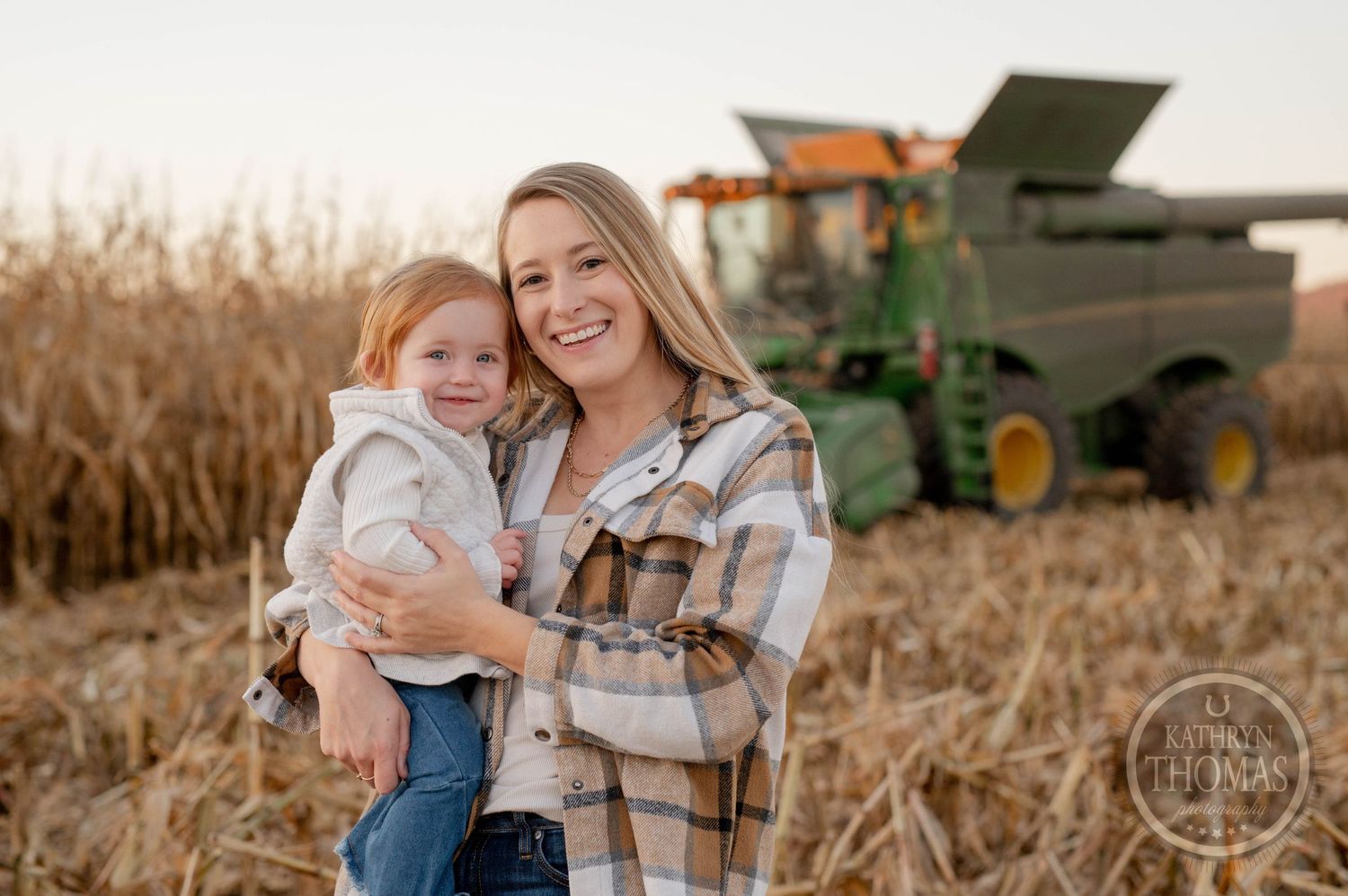 Mother and child smile together in cornfield with harvesting equipment in background.