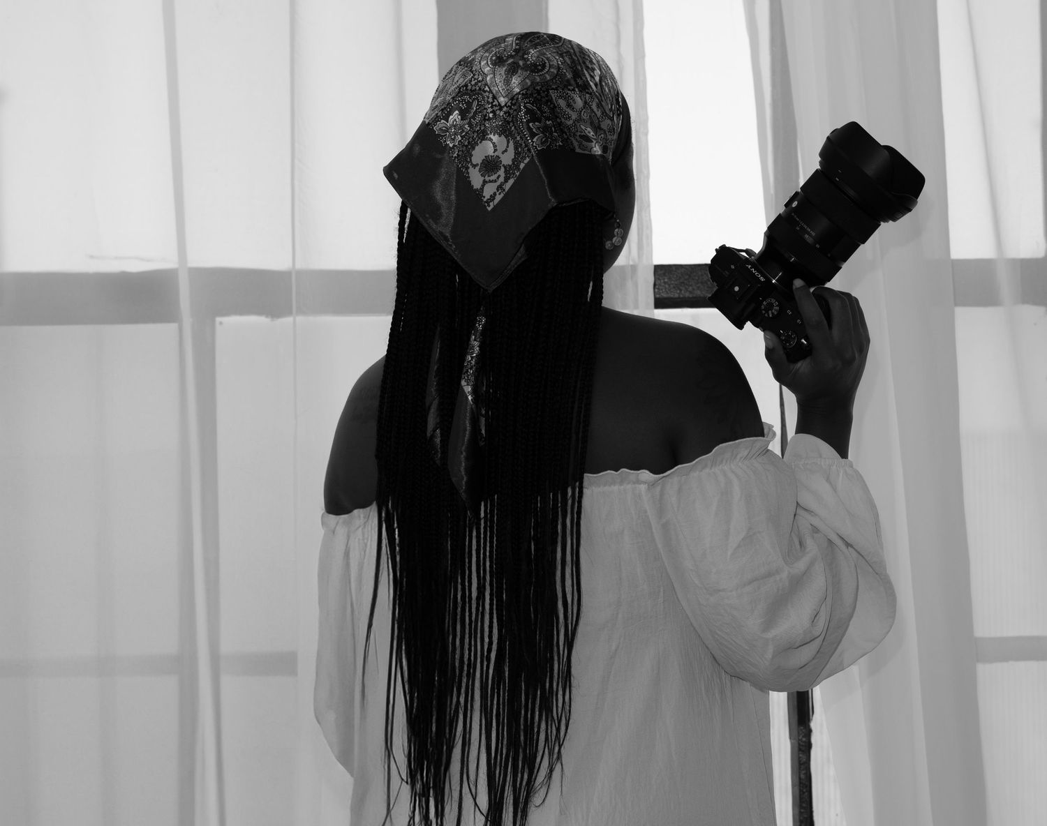 A black and white silhouette of someone with long dark hair holding a camera against sheer curtains.