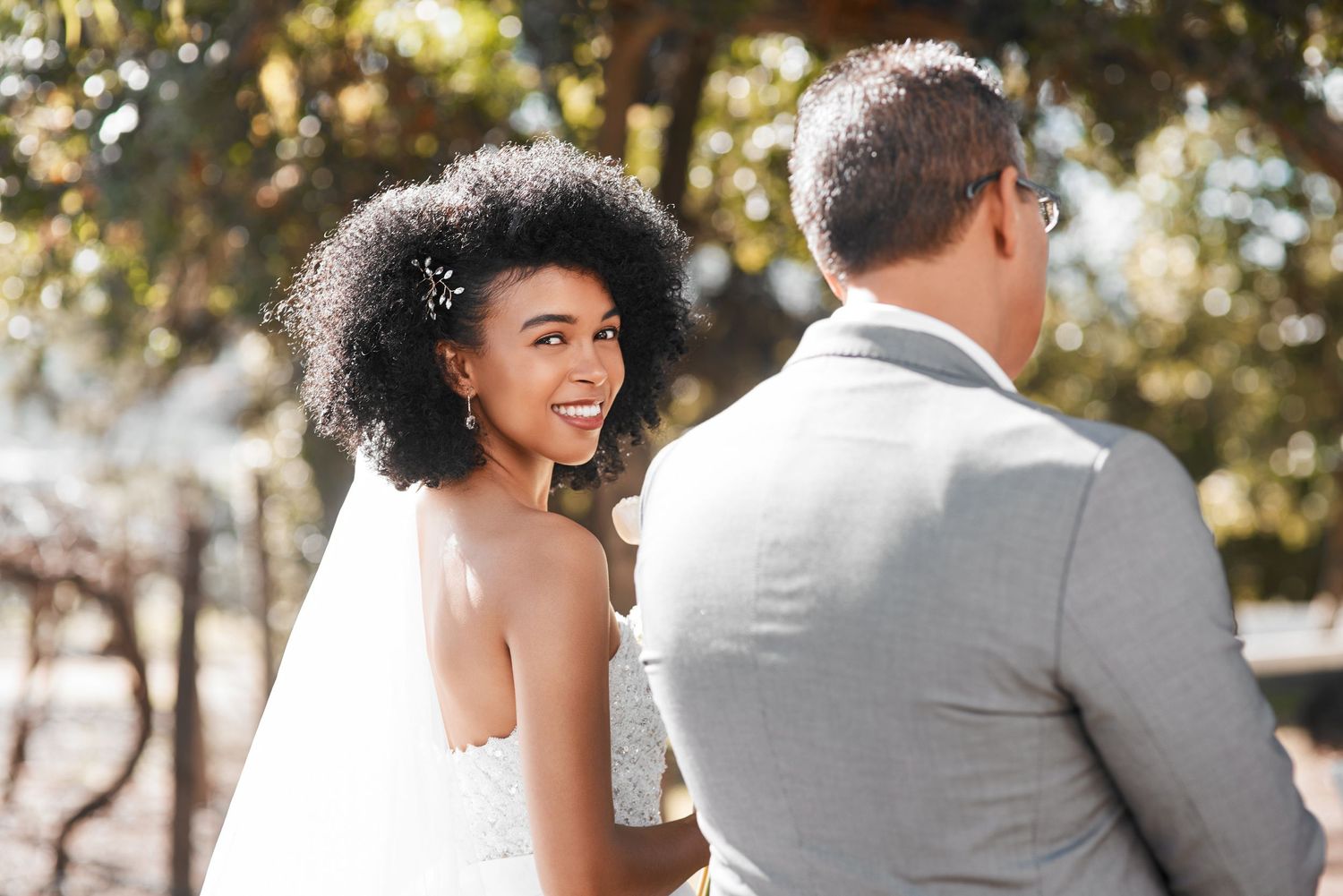 Bride with natural afro hairstyle smiles joyfully while standing next to her partner in a grey suit outdoors.