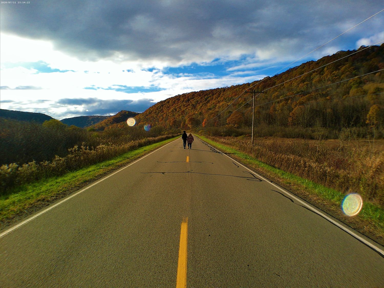 A lone figure walks down a winding mountain road surrounded by fall colors and cloudy skies.