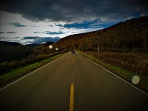A dramatic mountain road stretches into the distance under moody storm clouds and autumn hillsides.