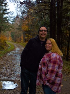 Two people stand together on a wet forest path surrounded by fall colors and evergreen trees.