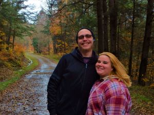 A couple poses on a rainy autumn trail with colorful fall foliage in the forest background.