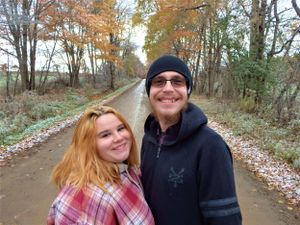 Two people smile together on a forest path with autumn trees in the background.