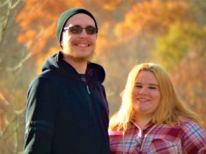 A couple poses outdoors against a backdrop of autumn foliage on a sunny fall day.