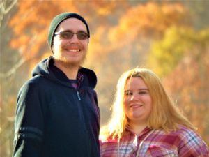 A couple smiles at the camera while standing together during a fall photoshoot.