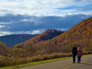 Two people walk along a mountain road surrounded by vibrant fall foliage and dramatic clouds.