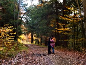 A couple embraces on a leaf-covered forest trail surrounded by fall colors.