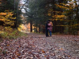 Two people share an intimate moment on a forest path covered in fallen autumn leaves.