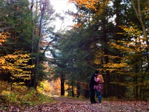 A couple stands together on a wooded trail surrounded by golden fall foliage.