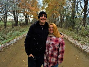 Two people share a tender moment on a tree-lined autumn path with fallen leaves.