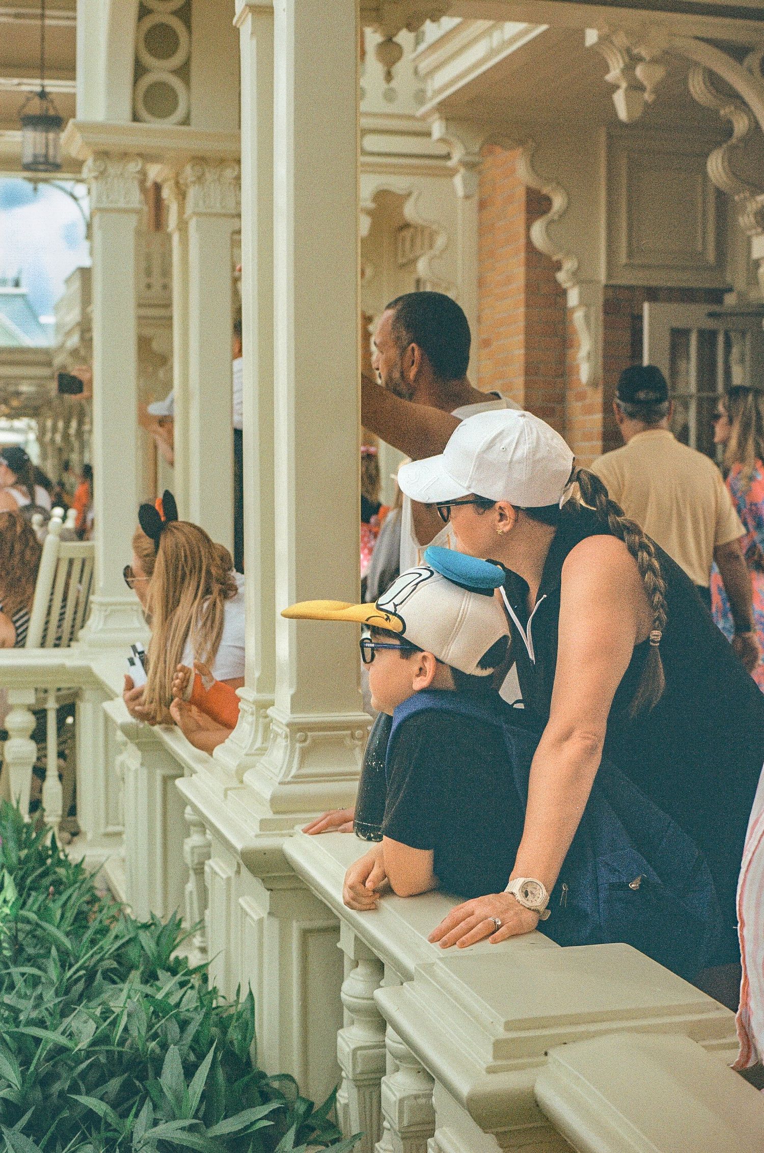 Visitors lean on an ornate white railing at a theme park, surrounded by decorative Victorian architecture and lush greenery.