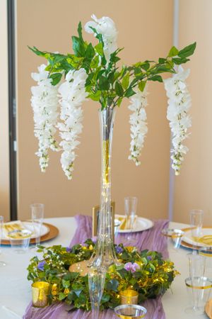 Elegant white wisteria centerpiece in tall glass vase with purple table runner and place settings.